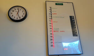 På en vegg i stuen henger en klokke med tydelige tall og en kalender som er delvis digital men også til å skrive på for hånd