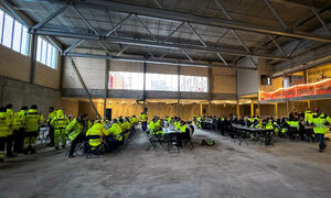 Arbeiderne i gule jakker sitter langs fire langbord i en stor hall