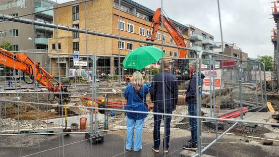 En dame med grønn paraply og to menn ser på gravemaskinen som graver i Ås sentrum
