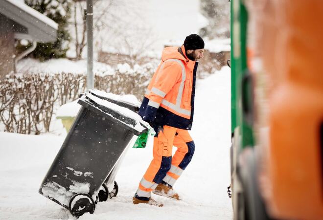 En mann i arbeidsklær drar søppeldunken mot søppelbilen i snøen