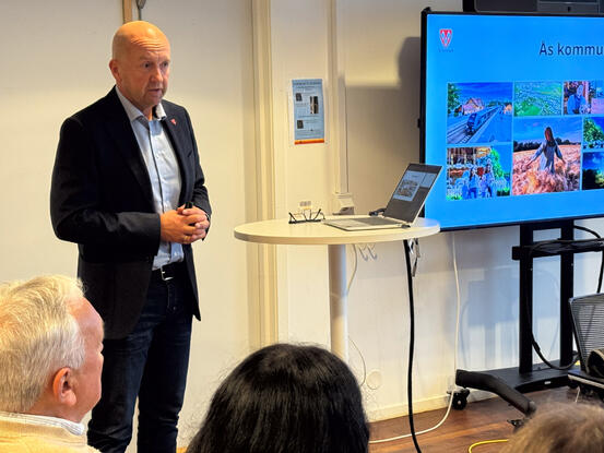 Kommunedirektør Tom-Arne Tørfoss la fram forslag til handlingsprogram med budsjett for formannskapet. Kommunedirektør Tom-Arne Tørfoss står foran en TV som viser bilder fra Ås