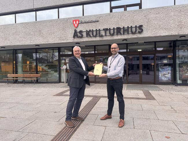 Kultursjef Kim Stian Gjerdingen Bakke (til høyre) overleverer temaplanen til Nils Sopp som leder hovedutvalg for oppvekst og kultur. Temaplanen overleveres utenfor Ås kulturhus