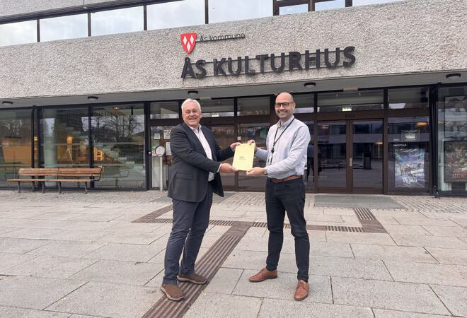 Kultursjef Kim Stian Gjerdingen Bakke (til høyre) overleverer temaplanen til Nils Sopp som leder hovedutvalg for oppvekst og kultur. Temaplanen overleveres utenfor Ås kulturhus